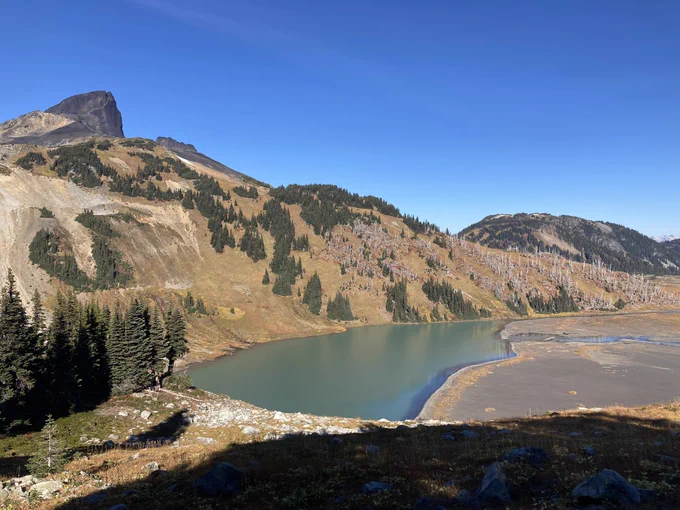 A photo of a blue alpine lake and it’s stream outlets. On the left side of the lake a mountain rises in the background, covered with patches of dark green conifers. Otherwise, the autumn vegetation is golden yellow.