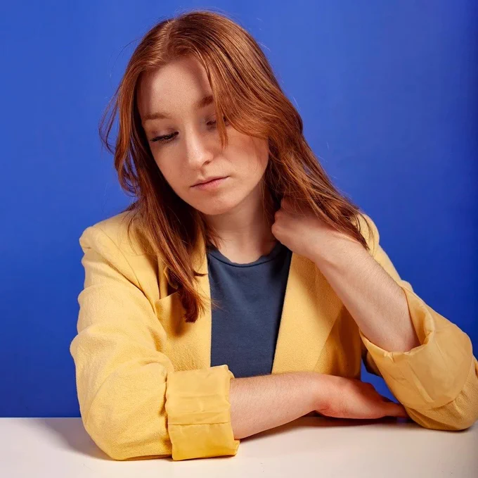  Kitty, a young white woman with long red hair, wearing a yellow jacket. She is seated and looking down at a table, with a bright blue background