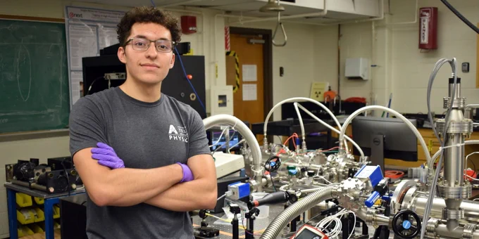 Photo of Javier Samaniego (a Hispanic man in a gray t-shirt and lab gloves) standing in a classroom in front of equipment used in a neutrino experiment that involves a lot of metal pumps and tubes