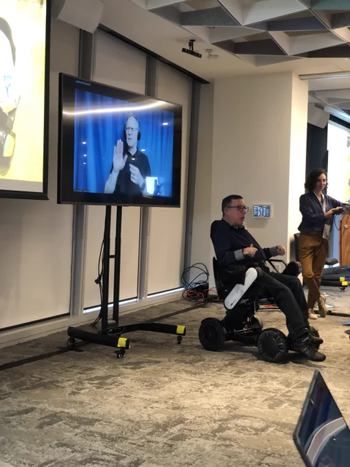 ALT TEXT: David Dume presenting at a11yTO sitting in his wheelchair in front of a screen with an ASL interpreter. 
