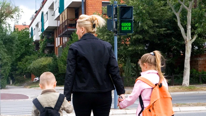 An adult woman crossing the street with two young children in front of a cross walk sign that reads "good luck."