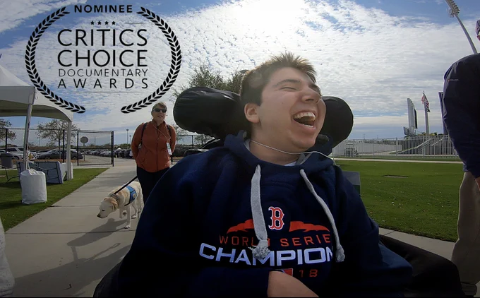 Photo of Samuel in his wheelchair, laughing. He's wearing a blue Red Sox sweatshirt. Behind him, Betsy is walking Proton. There is a bright blue sky with clouds. In the top left corner, black text reads "Nominee, Critics Choice Documentary Awards"