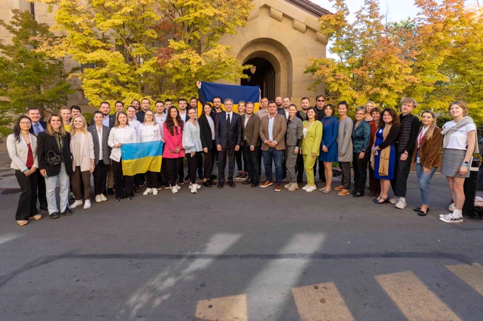 A large group of men and women, who are Silicon Valley startup founders from Ukraine, stand in front of a golden standstone building which is surrounded by green trees at Stanford University. Secretary Blinken stands at the center of the group. A Ukrainian flag hangs behind them. Two women also hold a smaller Ukrainian flag in the front row to the left of the group.