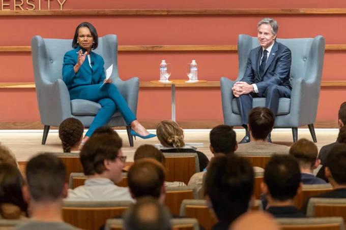 Former Secretary of State Condoleezza Rice and current Secretary of State Antony Blinken sit together on a raised stage in front of an audience at the Hoover Institute. Secretary Rice, wearing a teal suit, gestures with her hand, and holds a crisp piece of paper in the other while Secretary Blinken rests his hands on his knee and smiles. They both are seated in ice blue wing back chairs with a small table in between them that holds two clear bottles of water. The stage background is terra-cotta colored with horizontal strips of natural oak wood paneling. The audience members' heads are artfully out of focus in the foreground of the photo.