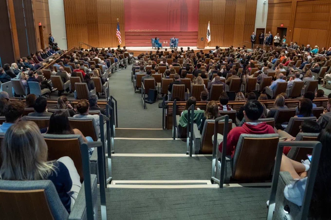 A wide angle camera view from the back of an auditorium, looking down at the large audience. Far, in the background of the photo, are former Secretary of State Condoleezza Rice and current Secretary of State Antony Blinken, sitting together on a raised stage. Secretary Rice, wearing a teal suit, gestures with her hand, and holds a crisp piece of paper in the other while Secretary Blinken rests his hands on his knee and smiles. Both are seated in ice blue wing back chairs. The stage background is terra-cotta colored with horizontal strips of natural oak wood paneling and to the sides are a U.S. flag and a flag of California.
