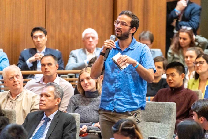 A man in a blue button down shirt and glasses, holding a pad of paper, stands with a microphone asking a question. Around him are fellow audience members, looking at him and listening.