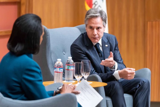Secretary of State Antony Blinken sits in a dark suit, gesturing with his hand and leaning in, in discussion, toward Former Secretary of State Condoleezza Rice. Secretary Rice, wearing a teal suit, holds a pen and crisp piece of paper in her hand while listening. They both are seated in ice blue wing back chairs with a small table in between them that holds two clear bottles of water and glasses. 