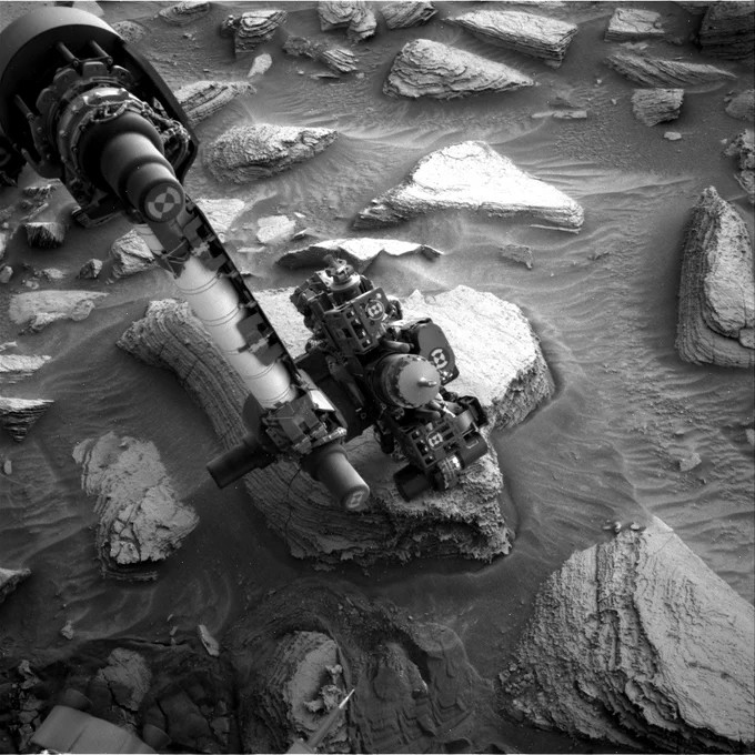 This black and white Navcam image, taken on sol 3624 (October 16, 2022), shows Curiosity's robotic arm placed over the recent drill hole named "Canaima". The light-toned block of layered rock is one among many in the area, separated by darker sand which forms centimeter-scale ripples.  Credit: NASA/JPL-Caltech
