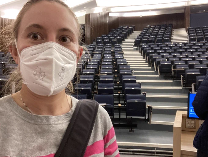 A photo of a woman in an N95 mask taking a selfie in front of a large empty lecture hall