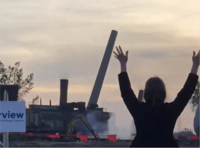 A woman with celebratory hands in the air as the smokestacks of a coke refinery are demolished