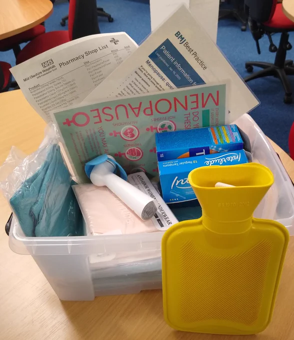 Box filled with sanitary products, a yellow hot water bottle, hand-held fan, underwear in different sizes, scrub trousers and information sheets on the Menopause from BMJ Best Practice, Pausitivity and the Pharmacy shop list.