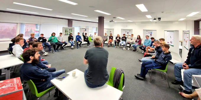A photograph of a large group of young people sat in a circle on lime green chairs. They are all listening to a man in a dark coloured t-shirt, who has his back to the camera. He is John Collison, co-founder of Stripe. He is speaking with University of Limerick students in a large white room