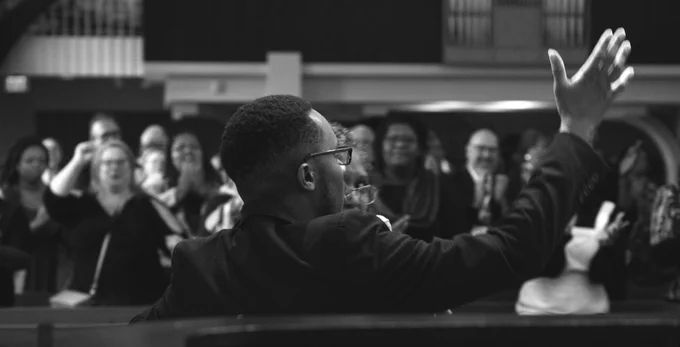 A Black musician faces a crowd with his back to the camera. He has his arm raised as he conducts a song. The crowd is blurred but singing along with the musician.