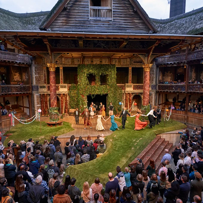 A green grass turf covers the wooden thrust of the Globe Theatre stage, an additional catwalk extending into the Yard (which is full of audience members). Actors wearing evening gowns and black tie suits hold hands, and dance around the stage, forming a chain. Climbing ivy covers the back wall of the theatre.