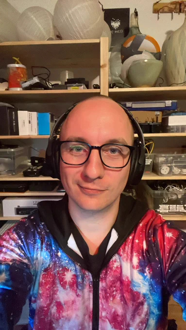Selfie of Tim in his home office, wearing a space onesie and wearing his headphones. A shelf of tools and electronics behind him.