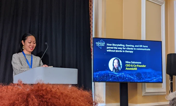 An Asian woman with red lipstick standing at a lectern. A slide next to her says "Nina Salomons, CEO and Co-founder, AnomieXR"