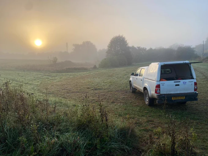 Misty green field with white work van parked on one side and sun shining through the must above.