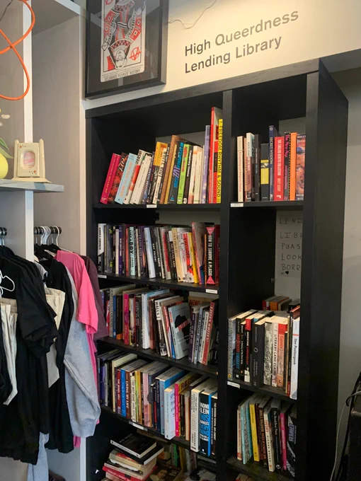 An image of a black book shelf filled with colourful, well-loved books. A sign above reads: High Queerdness Lending Library.