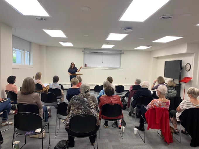In this photo, 2022-2023 Canadian Writer-in-Residence Leah Horlick stands at a podium at the front of a room. Audience members sit in black chairs, with rapt attention. 