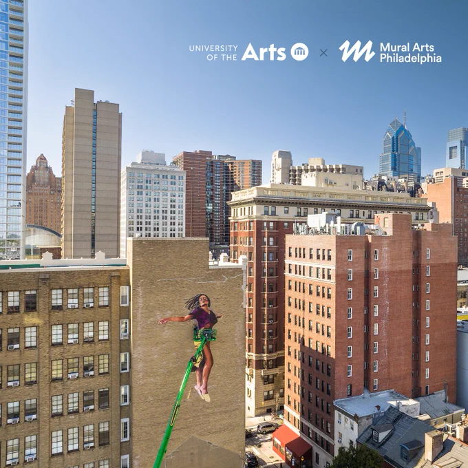 A series of city buildings, one with a mural of a person flying in the air. There is white text in the sky portion of the photo that reads "University of the Arts x Mural Arts Philadelphia"