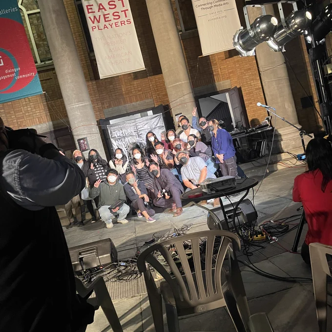 A slightly tilted photo of some of our TNC staff and performers in the courtyard of the Union Center for the Arts during our October 18th closer show. Everyone is masked up and smiling and some folks flash peace signs. A couple of folks are blocked out by an audience member in the immediate left foreground whoops. They’re all brightly lit by our stage lights.