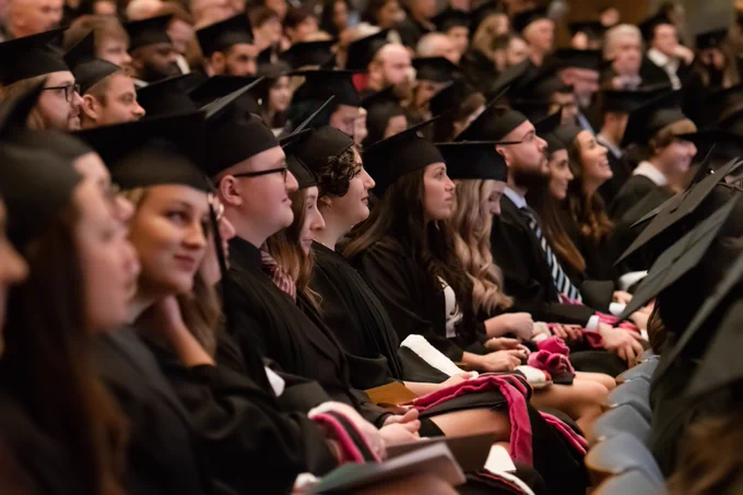 Graduates sit in rows in an auditorium wearing mortarboards and robes.