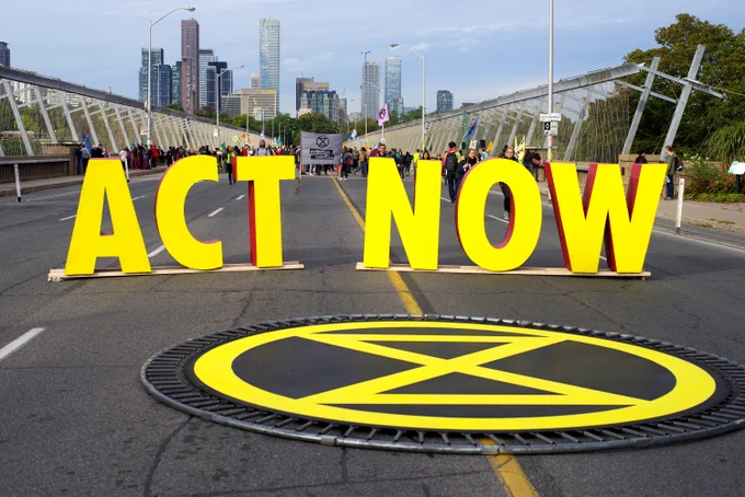 Huge 3D letters standing upright on a street spell out the words [ACT NOW], in front of which is the Extinction Rebellion symbol.