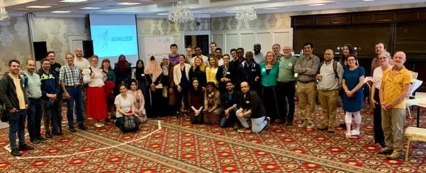 A picture of the Summer School attendees comprising trainers, supervisors, administrators and students. Everyone is smiling, the setting is the large, brightly lit and colourful conference room at the Glenroyal Hotel in Maynooth. There is a large projector screen in the background featuring the words: ADVANCE CRT.