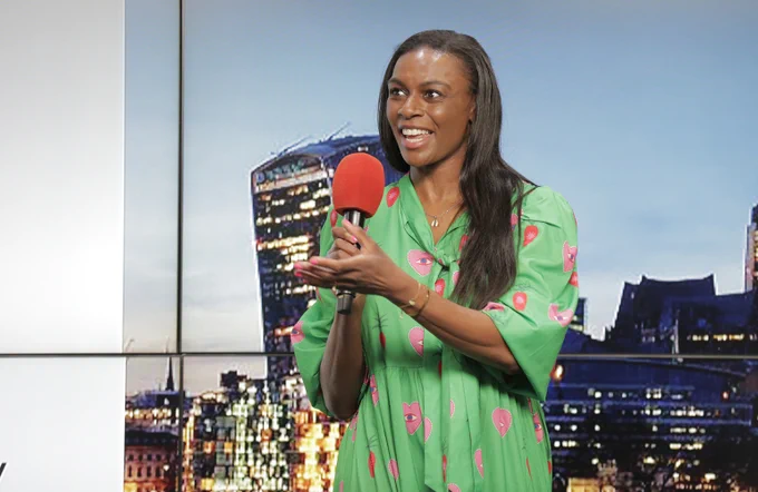 Camille speaking into a red microphone, smiling with her hand raised, wearing a green and pink outfit. 
