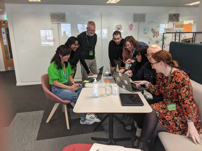Eu, Richa, Martin, Andy, Bev, Charlie, Katherine and Emma all stood and sat around laptops, deep in thought and conversation, looking and pointing at each others screens - but also laughing and smiling, because it's a staged photo!