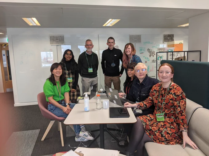 Eu, Richa, Martin, Andy, Beverley, Charlie, Katherine and Emma stood and sat around a table, smiling at the camera