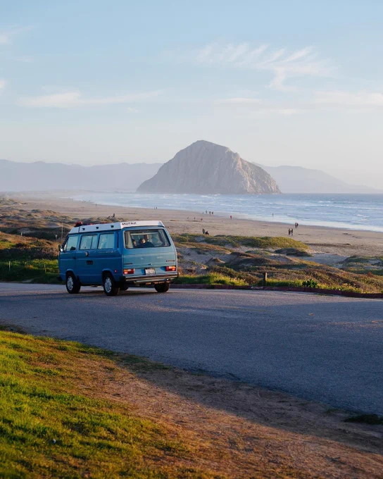 An old Westfalia driving down Highway 1 with a beach and giant rock in the distance