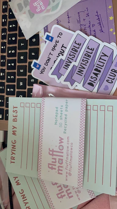 Photo of two "trying my best" notepads from @fluffmallow and a pile of stickers related to invisible disability.