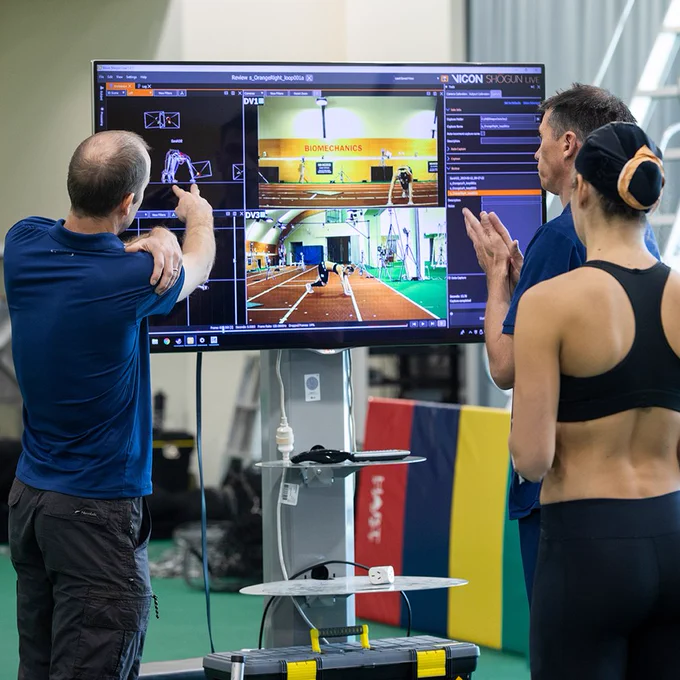Image shows three people looking at a large tv screen displaying data and vision of the athlete training. 