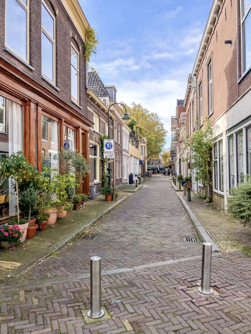 A red brick residential street in the center of Delft is lined with three-storey houses, planter boxes, and lighting fixtures. The front of the car-free street is blocked by two metal bollards.
