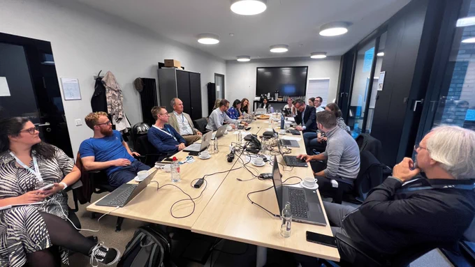 The picture shows several people gathering around a table. On the table, items like computers, bottles or papers can be seen. There is also a big screen at the back of the room. 