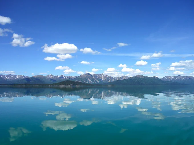 Lake Clark reflects the clouds and blue skies of an Alaska summer. In the background, a snow capped mountain range can be seen. Credit: NPS, Public Domain