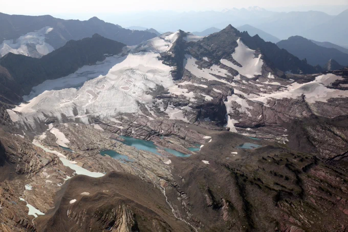Aerial view of Sperry Glacier on mountain. Small lakes and a mosaic of snow patches and bare mountain can be seen with mountain ridges in the background. 