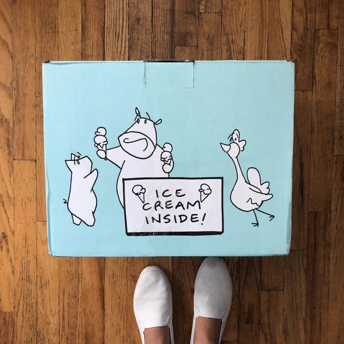 From above, A pair of feet in seamless shoes are seen before an Ample Hills shipping box that depicts PB The pig, Walt the Cow and Whitty the chicken next to a sign saying "Ice Cream inside!".]