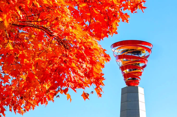 Red and orange tree canopy over a cyclone sculpture outside of the Alumni Center.