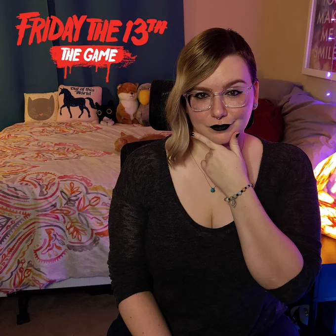 A picture of briteboom in dark clothes and makeup. There is a sleeping cat on the bed behind her and the logo for Friday the 13th: The Game is in the top left corner.