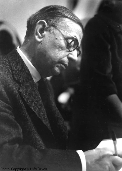 Black and white photo of Satre in profile. He is looking down and writing.