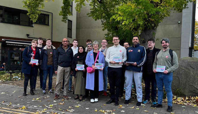Lilian and local councillor Pavlos are standing with a group of 13 other people outside the Avocafe on Derby Road, Nottingham, smiling and holding leaflets,