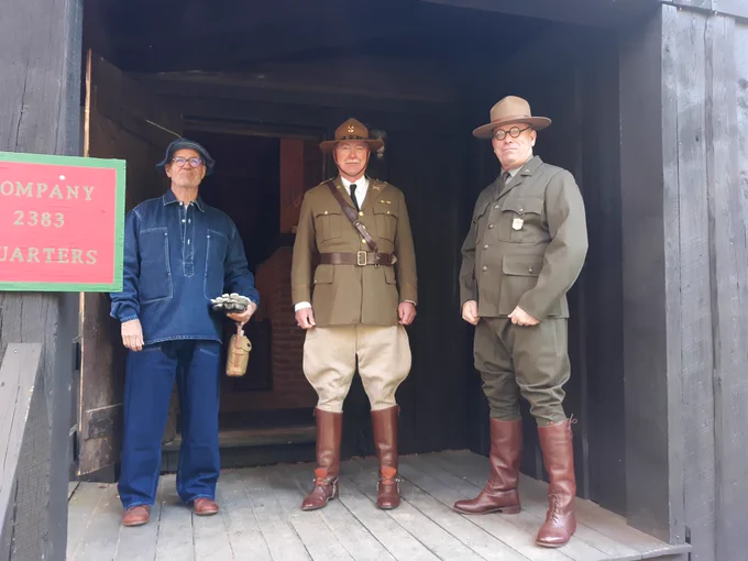 NPS Photo of three men from Company 2383 standing on a cabin porch modeling from the left, a blue CCC work uniform, the olive jacket, tan pants and high brown boots of the CCC dress uniform, and a ranger modeling a green NPS uniform from the 1930s