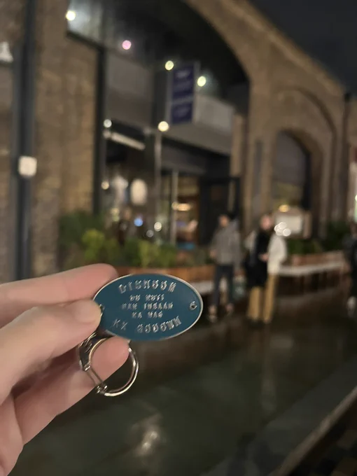 Green oval key chain with the name Dishoom engraved, with a restaurant is in the background