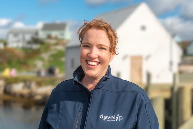 Leanne Jennings, Regional Lead for Project Planning and Delivery with Develop Nova Scotia, smiles at the camera in front of a Peggy's Cove scene with docks, water, and buildings in the background.
