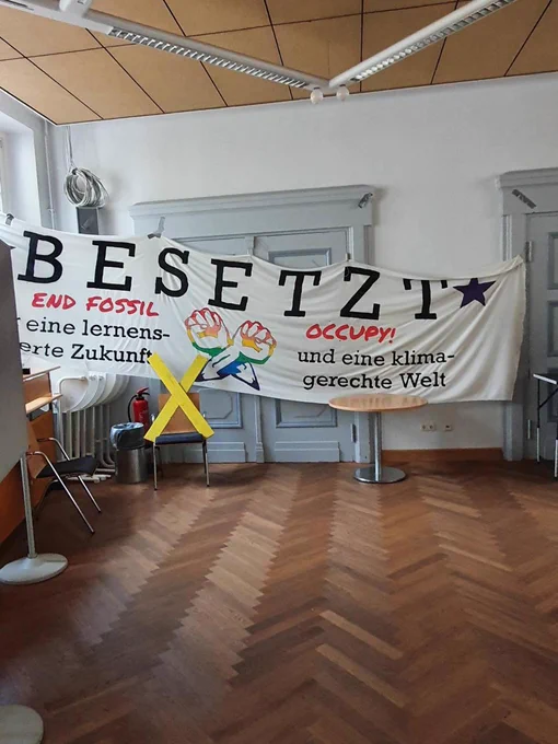 Ein weißes Banner mit der Aufschrift "Besetzt - End Fossil Occupy" vor einer weißen Tür in einer Schule. Davor Parkettfußboden