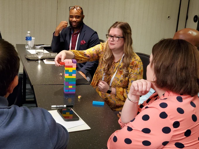 Woman attempting to pull a red block out of a multi-colored block tower while others look on. 