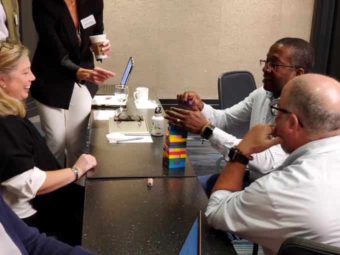 Man placing a block on top of a multi-colored block tower while others watch and smile. 