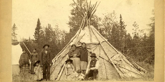 Family with children standing in or near a teepee made of poles, bark, and skins with a canvas tent nearby and trees behind them